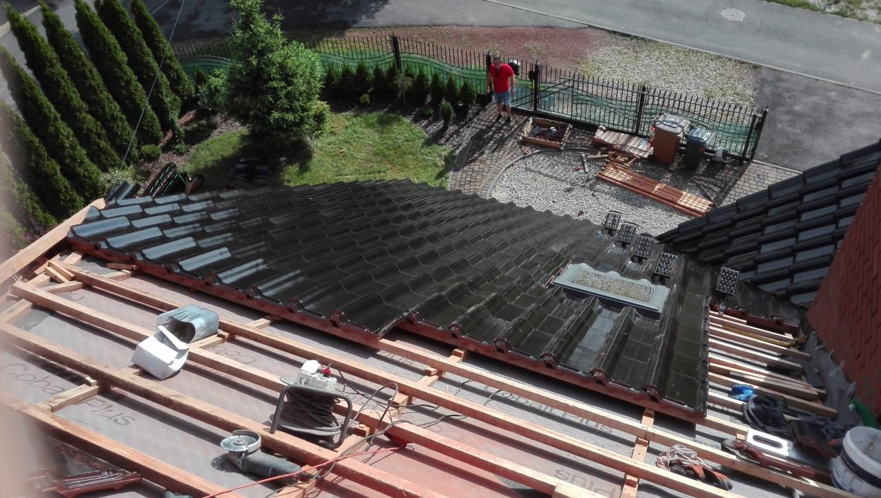 Częściowo pokryty dach z czarnej dachówki ceramicznej, widoczne drewniane łaty, membrana dachowa, narzędzia i pracownik w oddali.