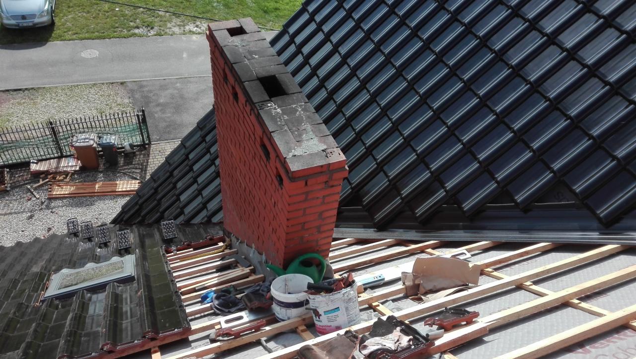Częściowy widok dachu w trakcie prac dekarskich, z widocznym kominem z czerwonej cegły, nową czarną dachówką i elementami konstrukcji drewnianej, narzędzia i puszki z farbą na dachu.