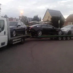 Laweta z trzema samochodami: ciemny BMW, czarny Jaguar i srebrny Citroen Picasso, zaparkowana na szarej asfaltowej drodze w tle domy z dachami pokrytymi panelami słonecznymi.