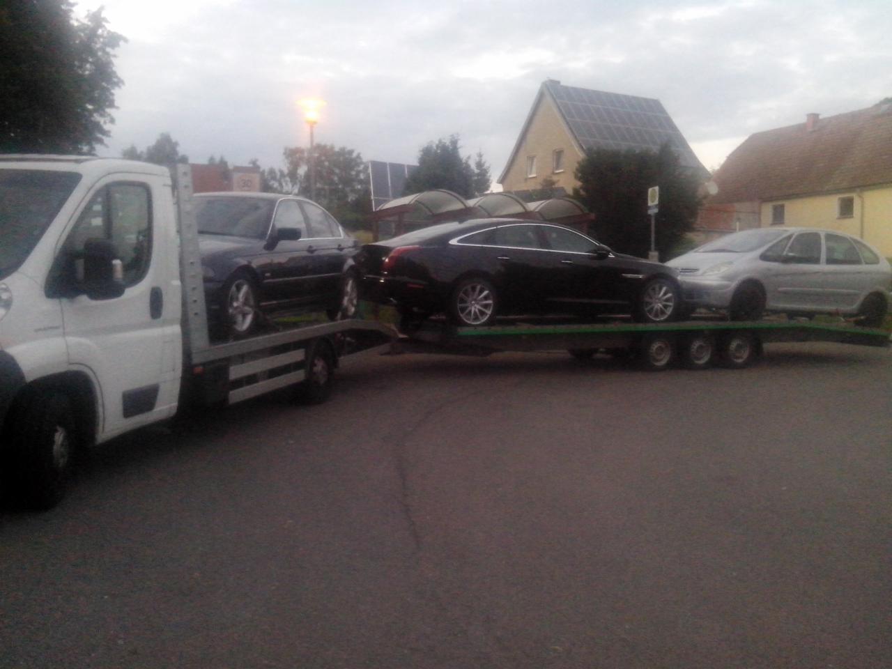 Laweta z trzema samochodami: ciemny BMW, czarny Jaguar i srebrny Citroen Picasso, zaparkowana na szarej asfaltowej drodze w tle domy z dachami pokrytymi panelami słonecznymi.