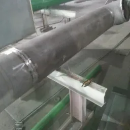 Fragment rury stalowej z widocznym spoiwem po spawaniu, umieszczonej na metalowych podporach w hali produkcyjnej.