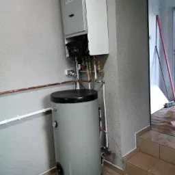 Kocioł gazowy kondensacyjny z podgrzewaczem wody, zamontowany na szarej ścianie w pomieszczeniu gospodarczym z płytkami na podłodze i schodami prowadzącymi do otwartego przejścia, widoczne rury...