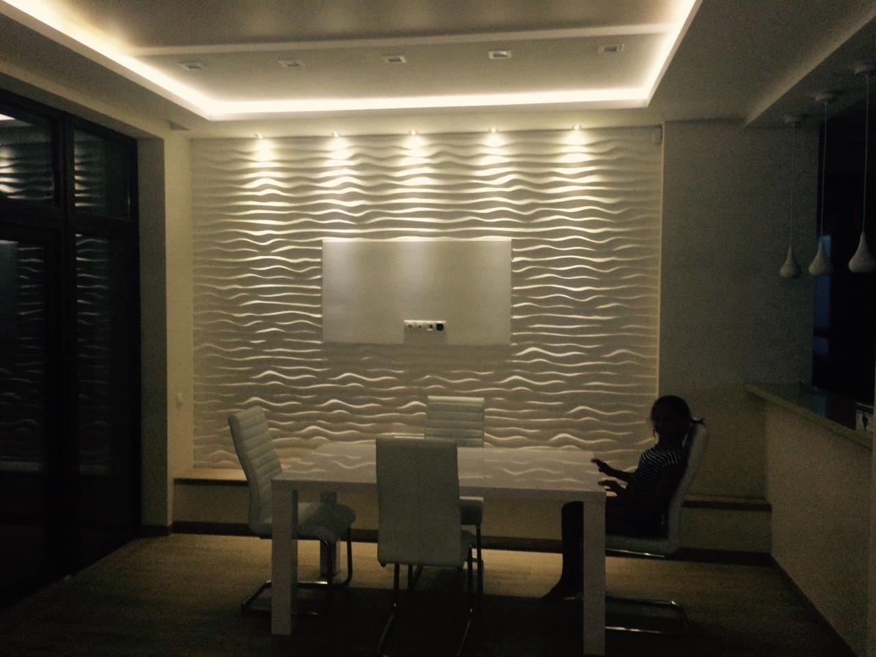 Nowoczesna jadalnia z białą ścianą 3D o falistym wzorze, oświetleniem LED i białym stołem z krzesłami; osoba siedząca przy stole.