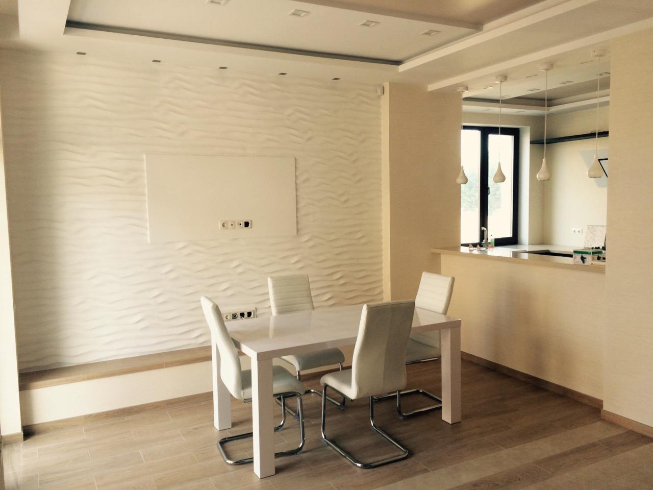 Nowoczesna jadalnia z białym stołem, krzesłami na chromowanych nogach i ścianą z dekoracyjną fakturą fal. Widoczna część kuchni z oknem i lampami.