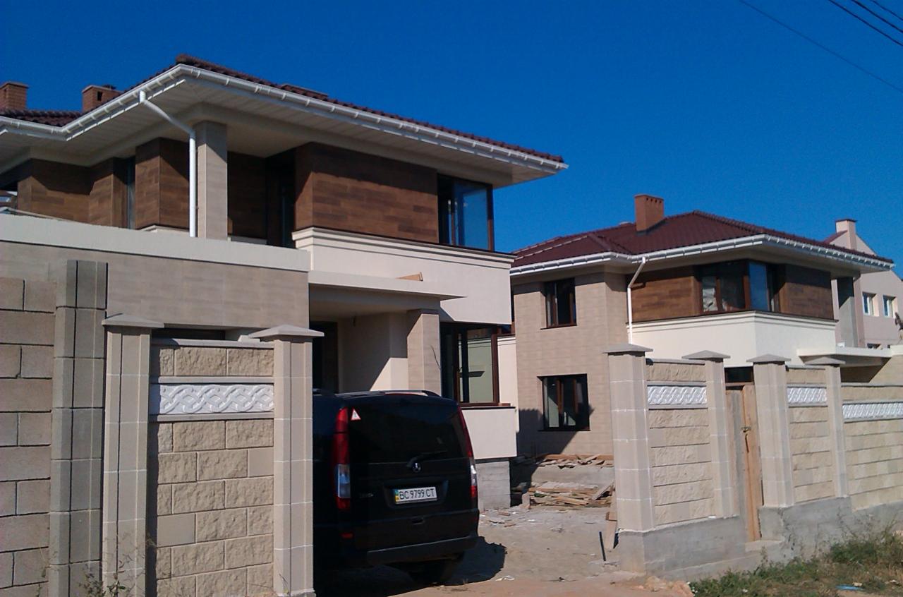 Dwa domy jednorodzinne z ogrodzeniem z kostki łupanej z dekoracyjnym elementem, na podjeździe zaparkowany czarny van.