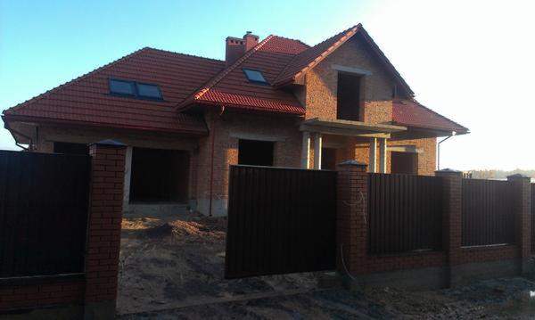 Niedokończony dom z czerwonej cegły z brązowym dachem i ciemnobrązowym ogrodzeniem panelowym z ceglanymi słupkami w słoneczny dzień.