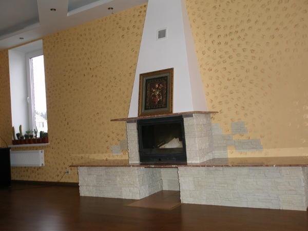 Wykończony kominek z obudową z jasnej imitacji kamienia, z brązowym blatem, na tle ściany z wzorzystą tapetą, z obrazem nad paleniskiem.