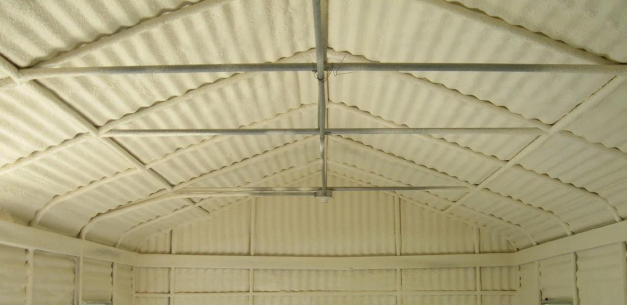 Wnętrze budynku z blachy falistej pokryte grubą warstwą pianki izolacyjnej, widoczne metalowe rury podtrzymujące konstrukcję dachu.