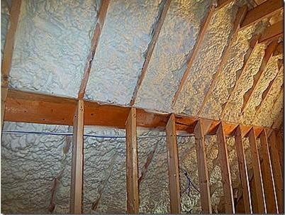 Poddasze z drewnianą konstrukcją pokryte jasną, nierówną warstwą izolacji natryskowej, widoczne przewody.