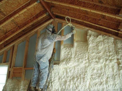 Pracownik w kombinezonie ochronnym aplikuje białą pianę izolacyjną na ścianę poddasza z drewnianą konstrukcją szkieletową, stojąc na drabinie.