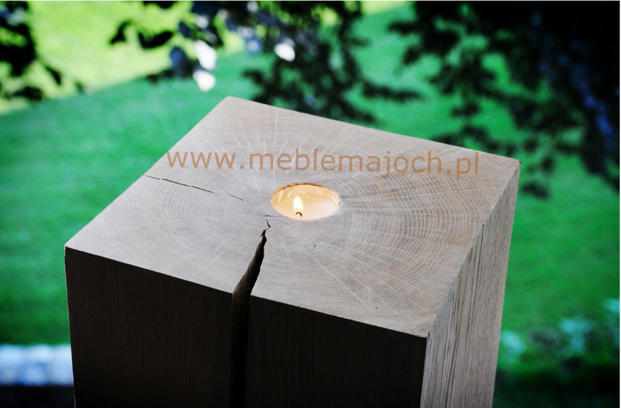 Kwadratowa, surowa, drewniana kolumna z widocznym pęknięciem i wywierconym otworem na świeczkę, na tle zieleni.