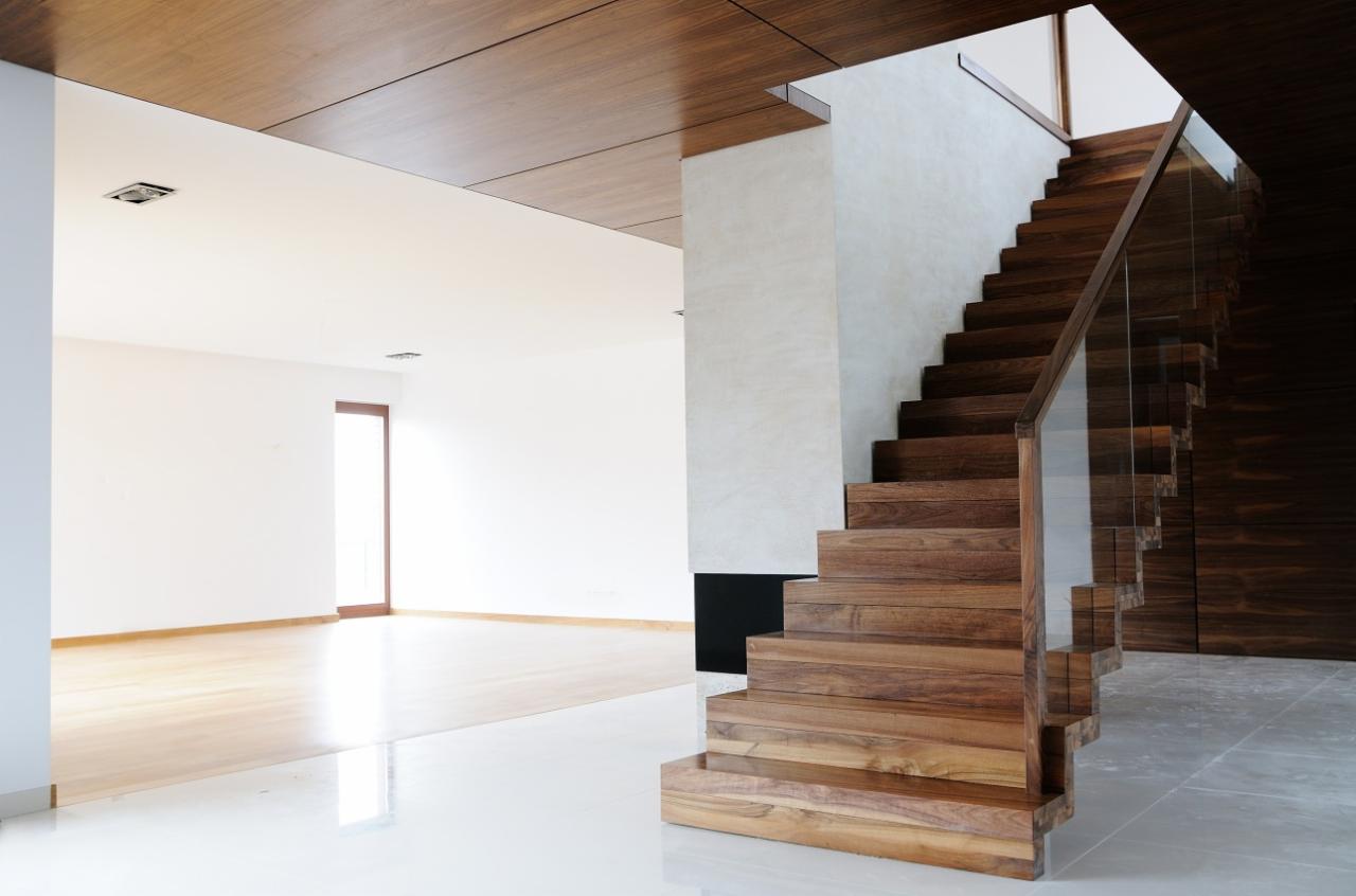 Nowoczesne schody z drewna egzotycznego z balustradą ze szkła hartowanego, widoczne z dołu, w tle jasne pomieszczenie z drewnianą podłogą i sufitem.