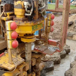 Ż&oacute;łta wiertnica Beretta w trakcie wykonywania przewiertu sterowanego, widoczne błoto i woda w wykopie, fragment rury w ziemi.