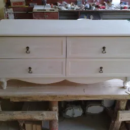 Niedokończona komoda z czterema szufladami, wykonana z jasnego drewna, z ozdobnymi uchwytami, stoi na prowizorycznym stole warsztatowym w trakcie montażu, widoczna wiertarka akumulatorowa obok.