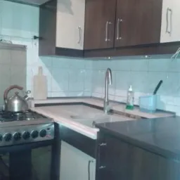 Fragment kuchni z zabudową: górne szafki w kolorze brązu i beżu, dolne szafki z beżowymi frontami, metalowy zlew i bateria, kuchenka gazowa z czajnikiem, białe kafelki na ścianie.