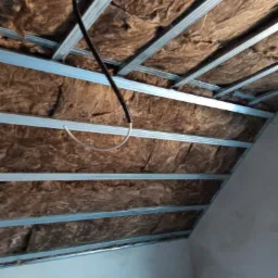 Skośne poddasze z widoczną konstrukcją metalową i brązową wełną mineralną, z czarnym kablem elektrycznym w peszlu oraz wystającym białym przewodem