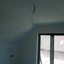 Poddasze w trakcie wykańczania, widoczne okno dachowe w sąsiednim budynku, kabel wystający z sufitu i przygotowane punkty elektryczne na ścianie.