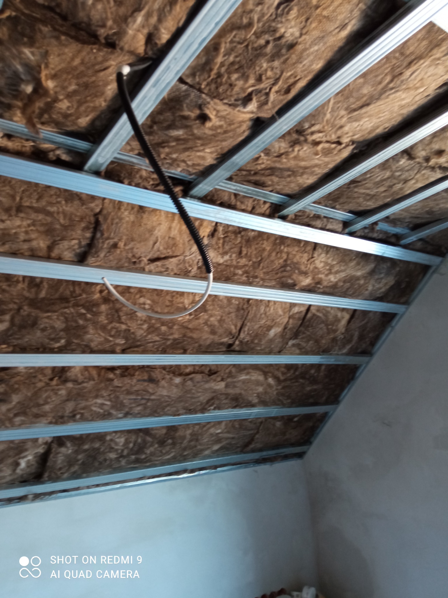 Skośne poddasze z widoczną konstrukcją metalową i brązową wełną mineralną, z czarnym kablem elektrycznym w peszlu oraz wystającym białym przewodem