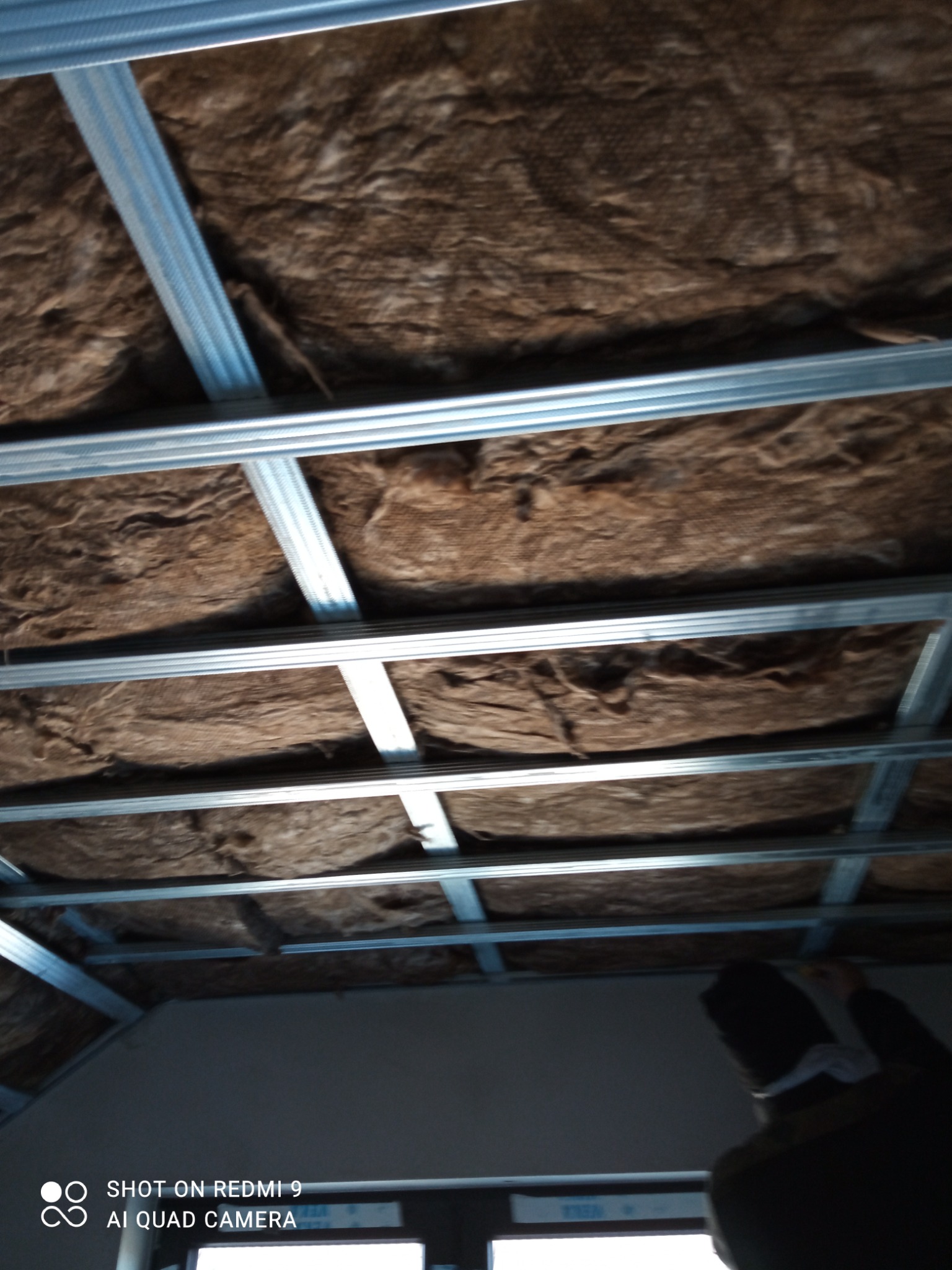 Montaż metalowej konstrukcji sufitu z wypełnieniem z wełny mineralnej, widok z dołu, fragment sylwetki pracownika w prawym dolnym rogu.