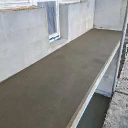 Świeżo wylana, gładka posadzka jastrychowa na balkonie, widoczne okna zabezpieczone folią, szare ściany w tle i fragment rusztowania.