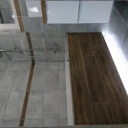 Wykończona łazienka z szarymi płytkami imitującymi kamień, drewnopodobnymi panelami w kabinie prysznicowej, oświetleniem LED wzdłuż podłogi i chromowaną armaturą.