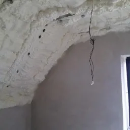 Poddasze z widoczną warstwą izolacyjną z pianki na skośnym suficie, z fragmentem okna w tle. Widoczne metalowe elementy konstrukcji pod pianką.