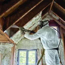 Aplikacja pianki PUR na poddaszu przez pracownika w kombinezonie ochronnym i masce, widoczne drewniane elementy konstrukcji dachu i okno.