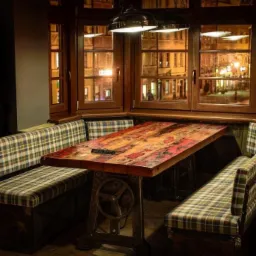 Rustykalny stół z blatem z odzyskanego drewna i metalową podstawą w stylu industrialnym, otoczony tapicerowanymi ławkami w kratę, ustawiony przy oknie z widokiem na nocne miasto.