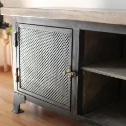 Komoda w stylu industrialnym z metalową siatką na drzwiczkach, drewnianym blatem i krótkimi, masywnymi nóżkami, na podłodze z paneli.