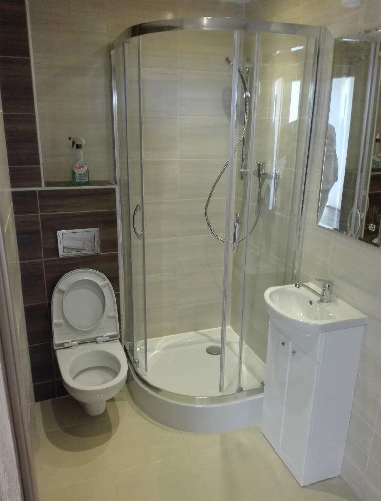 Zmodernizowana łazienka z narożną kabiną prysznicową, wiszącą toaletą z otwartą deską i białą szafką pod umywalką, uzupełniona beżowymi i brązowymi płytkami oraz butelką środka czyszczącego na półce.