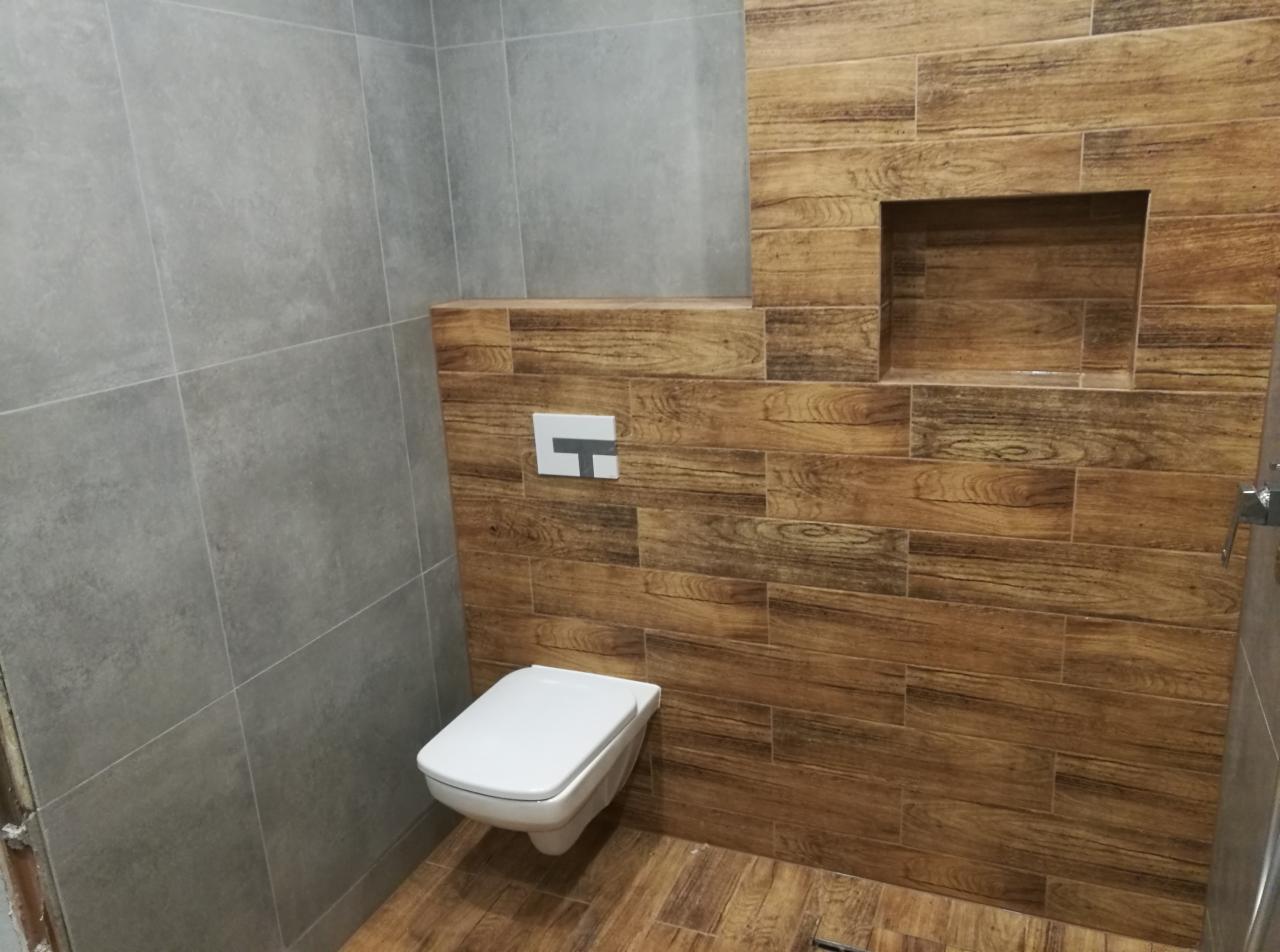 Nowoczesna łazienka z podwieszaną toaletą, szare płytki na jednej ścianie, drewnopodobne płytki na pozostałych, wnęka dekoracyjna w ścianie z drewnopodobnych płytek.