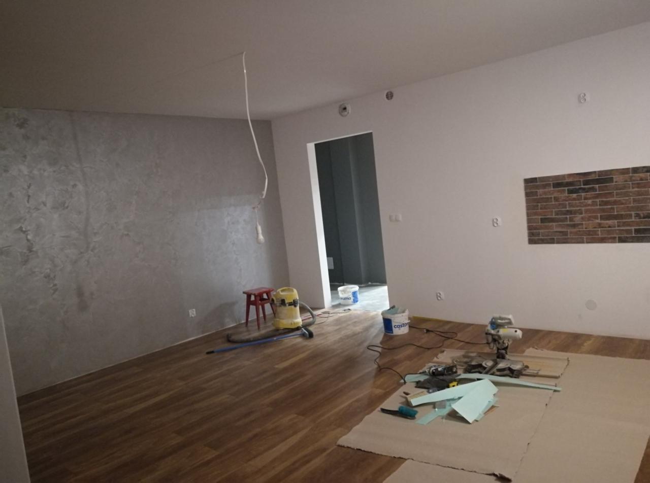 Pokój w trakcie prac wykończeniowych: położona podłoga z paneli drewnopodobnych, ściana z efektem dekoracyjnym, przygotowane narzędzia do cięcia paneli, odkurzacz i kubełki po farbie.