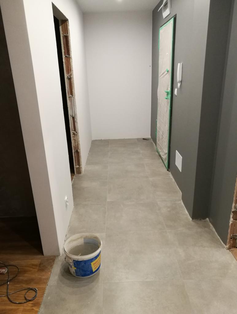 Korytarz w trakcie remontu: świeżo położone szare płytki podłogowe, ściany częściowo pomalowane na biało i szaro, drzwi zabezpieczone folią malarską, wiadro z wodą i narzędzia na podłodze.