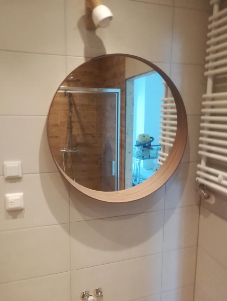 Okrągłe lustro z drewnianą ramą zawieszone na kafelkowanej ścianie w łazience, widoczne włączniki światła, grzejnik drabinkowy i prysznic z drewnopodobnymi płytkami w odbiciu.