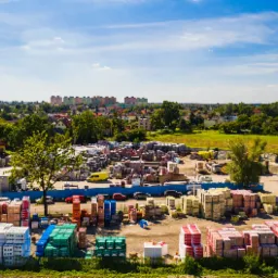Widok z góry na plac składowy materiałów budowlanych z ułożonymi paletami z cegłami, bloczkami i innymi materiałami, w tle zabudowa miejska z blokami i kościołem, błękitne niebo z chmurami.