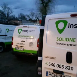 Flota trzech białych samochodów dostawczych marki Renault, z zielonym logo 'InstalExpert' i napisem 'Solidne Instalacje', zaparkowana na zewnątrz w pochmurny dzień, z widocznymi numerami telefonów...