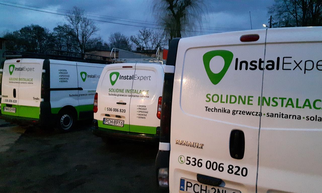 Flota trzech białych samochodów dostawczych marki Renault, z zielonym logo 'InstalExpert' i napisem 'Solidne Instalacje', zaparkowana na zewnątrz w pochmurny dzień, z widocznymi numerami telefonów...
