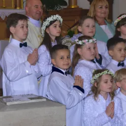 Grupa dzieci w białych strojach komunijnych z wiankami na głowach, stojących w kościele podczas uroczystości, z księdzem i rodzicami w tle. Uroczysty nastrój.