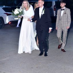 Zbigniew Płotka - e-foto-art - Para młoda w Warszawie, trzymająca się za ręce, w towarzystwie gości weselnych. Panna młoda w białej sukni i futrzanym etolu, pan młody w czarnym smokingu.