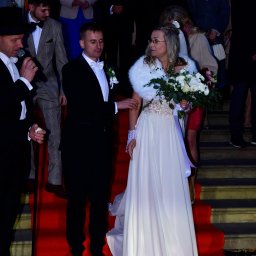 Zbigniew Płotka - e-foto-art - Para młoda na czerwonym dywanie, otoczona gośćmi. Panna młoda w białej sukni i futrzanym etolu, pan młody w smokingu. W tle schody i zgromadzeni ludzie.