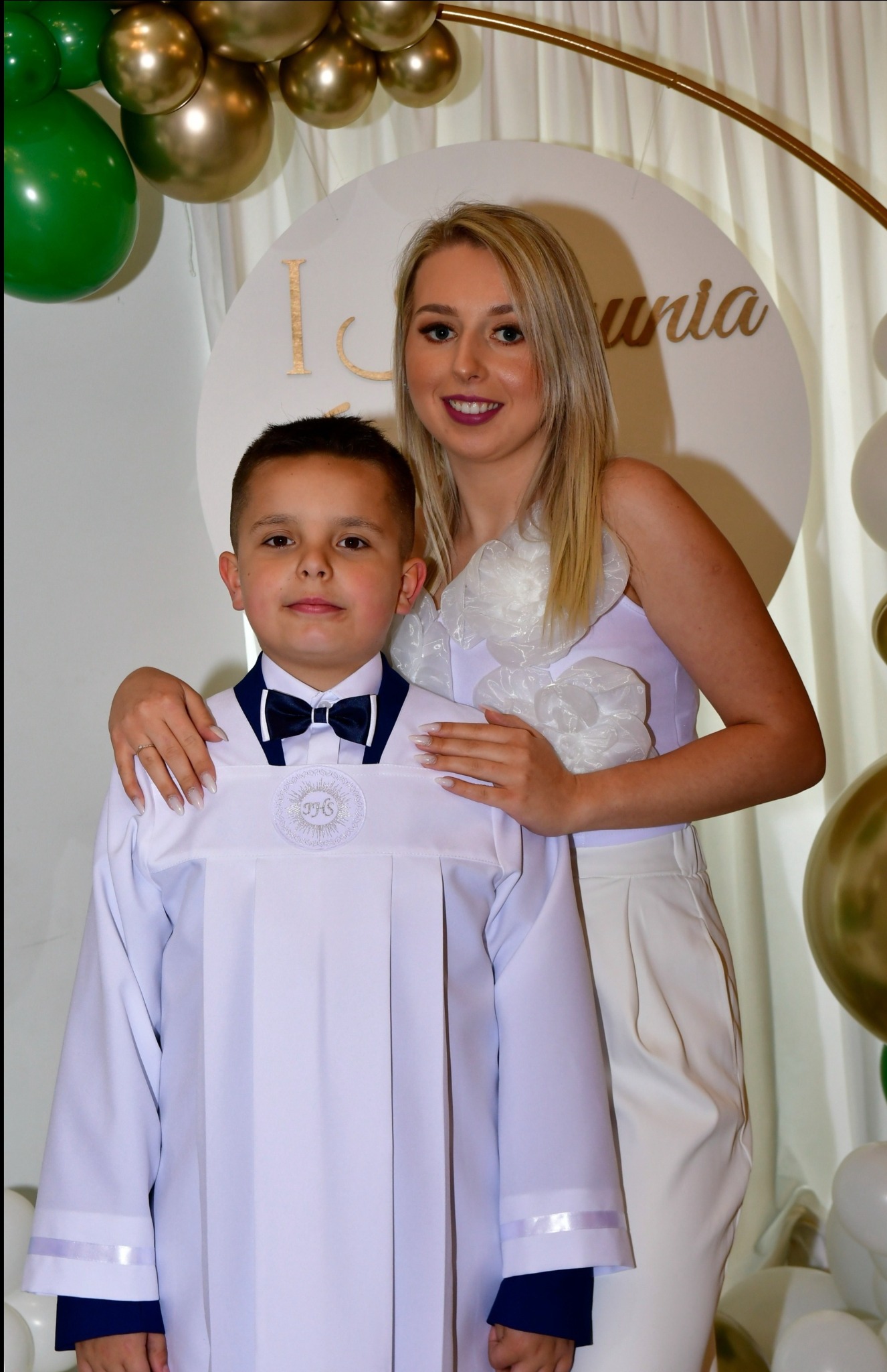 Chłopiec w albie komunijnej pozuje z mamą na tle dekoracji z balonów. Ujęcie portretowe, jasne kolory, subtelne oświetlenie.