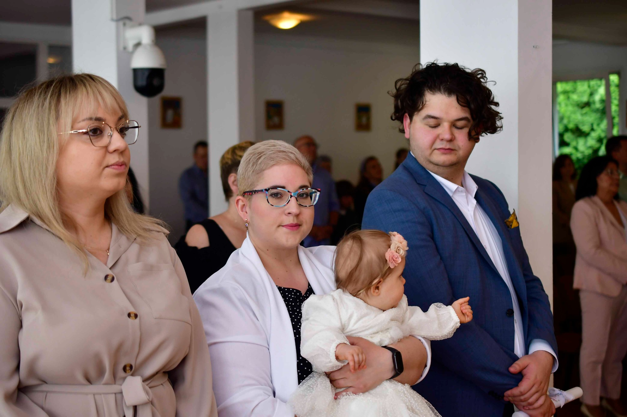 Uroczystość chrztu: rodzice trzymają ubraną na biało córeczkę w kościele, w tle widoczni goście i filar z kamerą monitoringu.