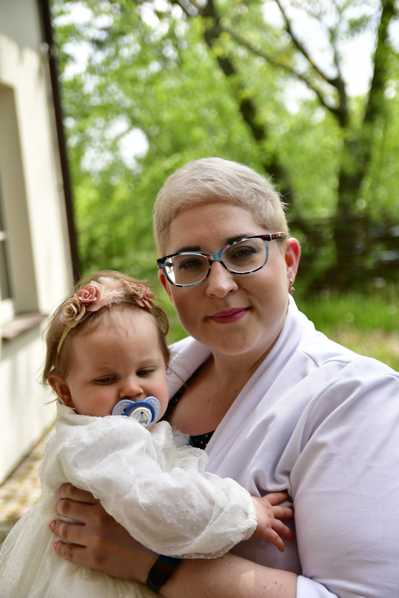 Mama w okularach trzyma na rękach niemowlę z smoczkiem i opaską z kwiatami. Zdjęcie w plenerze, rozmyte tło z zielenią.