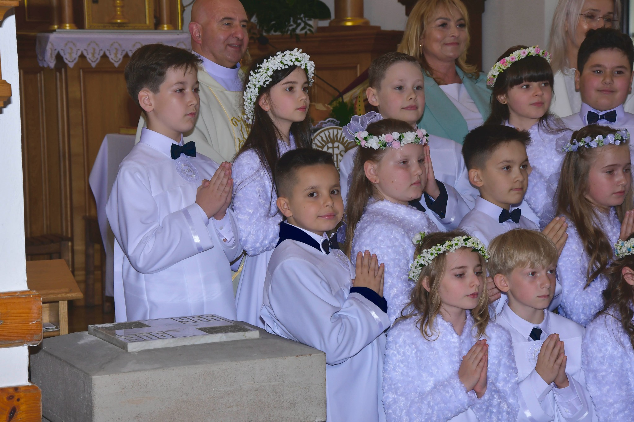 Grupa dzieci w białych strojach komunijnych z wiankami na głowach, stojących w kościele podczas uroczystości, z księdzem i rodzicami w tle. Uroczysty nastrój.
