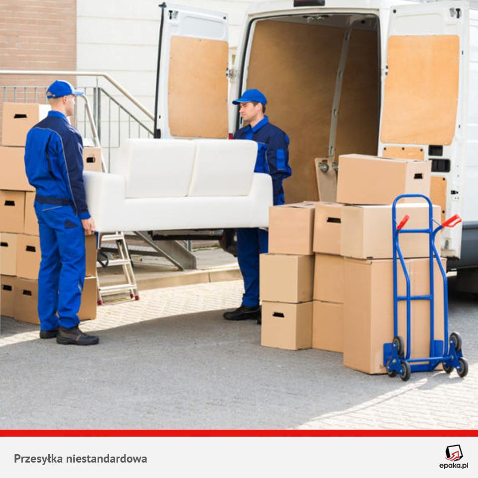 Dwóch pracowników w niebieskich kombinezonach wnosi białą sofę do białego busa, obok stoją kartony i niebieski wózek transportowy.