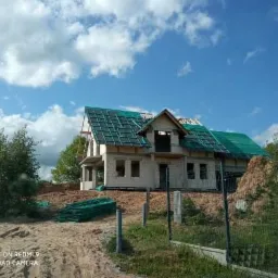 Budowa domu jednorodzinnego z białego bloczka, zabezpieczona zieloną siatką na dachu, widoczna konstrukcja więźby dachowej, teren budowy z piaskiem i zielenią w tle, pochmurne niebo.