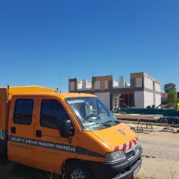 Pomarańczowy samochód dostawczy z napisem 'Projekty-Budowy-Nadzory-Geodezja' na tle budowy domu z białego pustaka, widoczni dwaj pracownicy w żółtych kamizelkach.