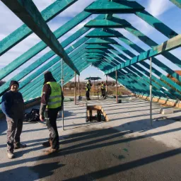 Konstrukcja drewnianego dachu w trakcie budowy, widoczni robotnicy w odblaskowych kamizelkach, niebieskie krokwie i więźba dachowa na tle błękitnego nieba.