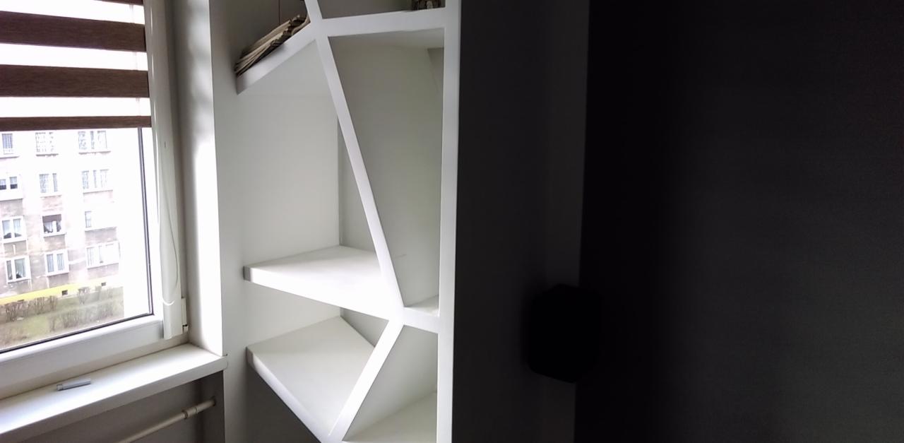 Biała, niestandardowa zabudowa regałowa z półkami, zlokalizowana obok okna z widokiem na budynek mieszkalny, na tle ciemnej ściany.