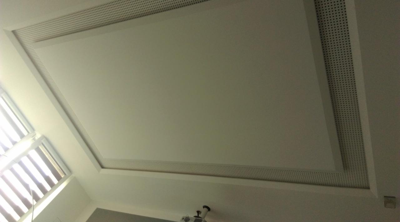 Biały sufit podwieszany z perforowanym obramowaniem, widok z dołu, oświetlenie dzienne z okna po lewej stronie.